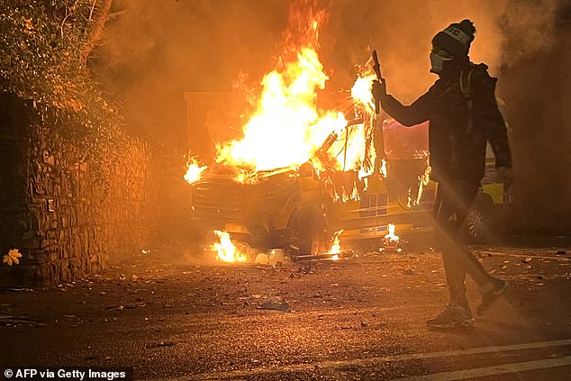 A protester films and a police vehicle burns in the background as a demonstration outside a hotel housing asylum seekers turns violent in Saggart, south-west of Dublin in Ireland on October 21, 2025